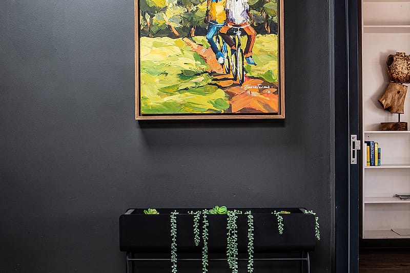 A framed painting of two cyclists on a country road, above a console of trailing plants.