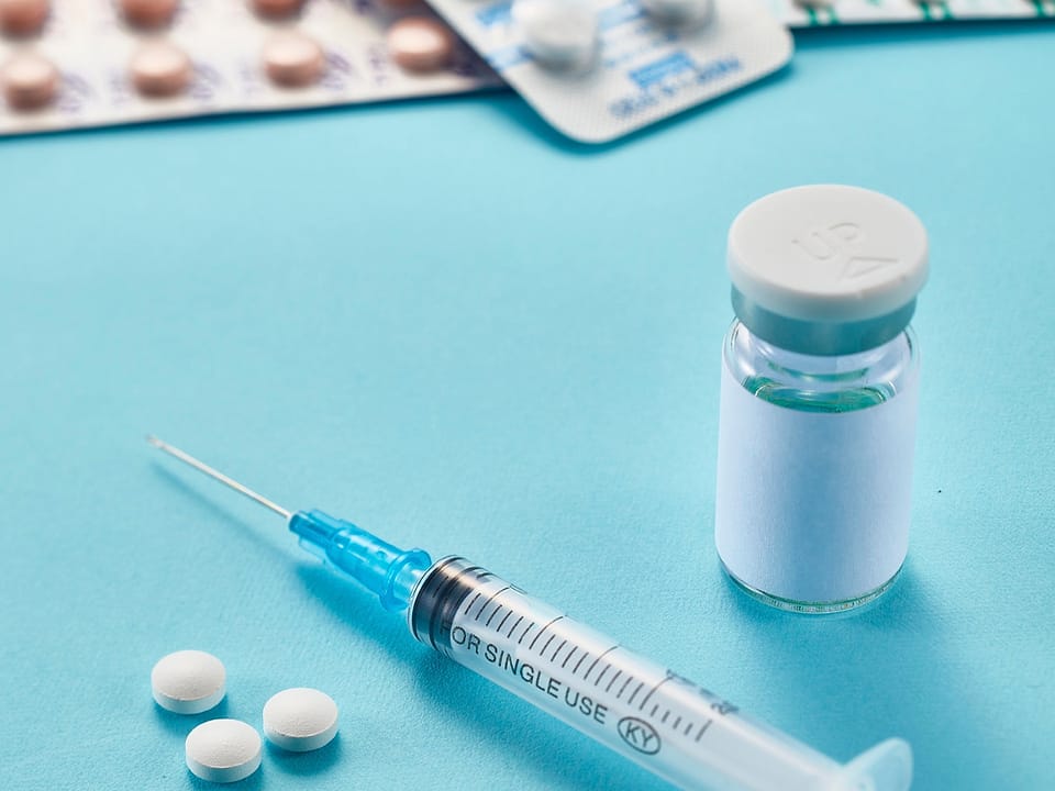 An injection pen, a glass vial and tablets laid out on a light blue background.