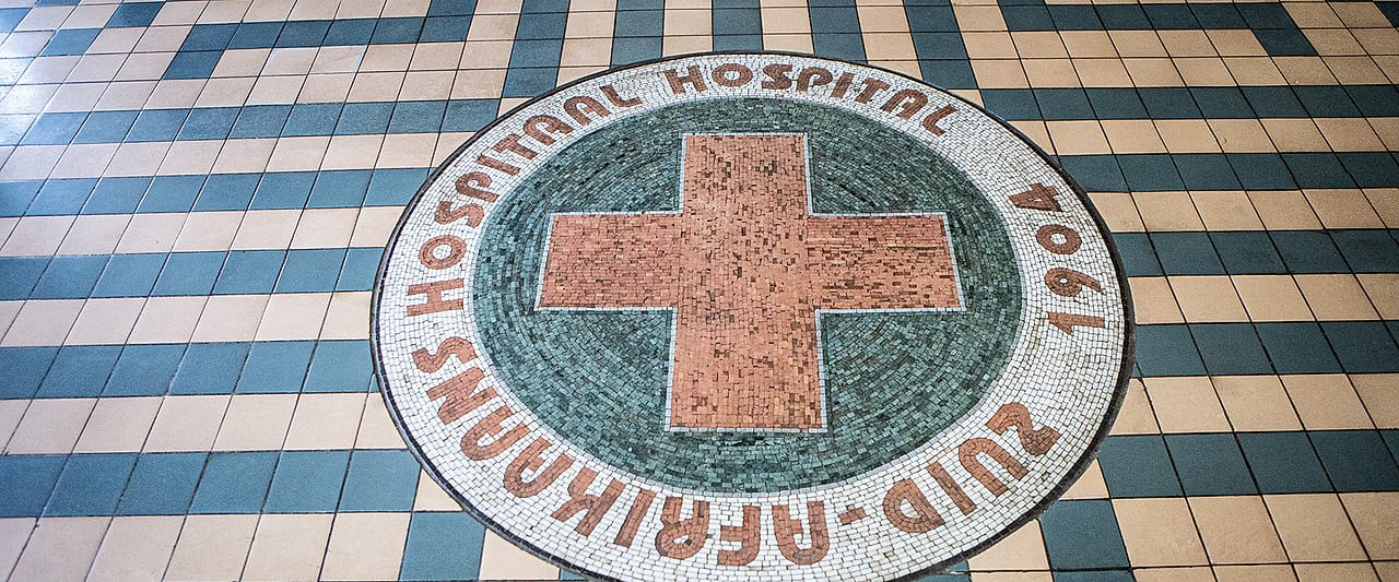 The original 1904 mosaic floor at Zuid-Afrikaans Hospital, a red cross set into tiled lettering reading “Zuid-Afrikaans Hospitaal”.